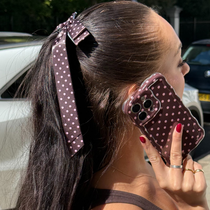 Woman wearing a brown polka dot ribbon bow hair clip holds a matching brown polka dot phone case to her ear.