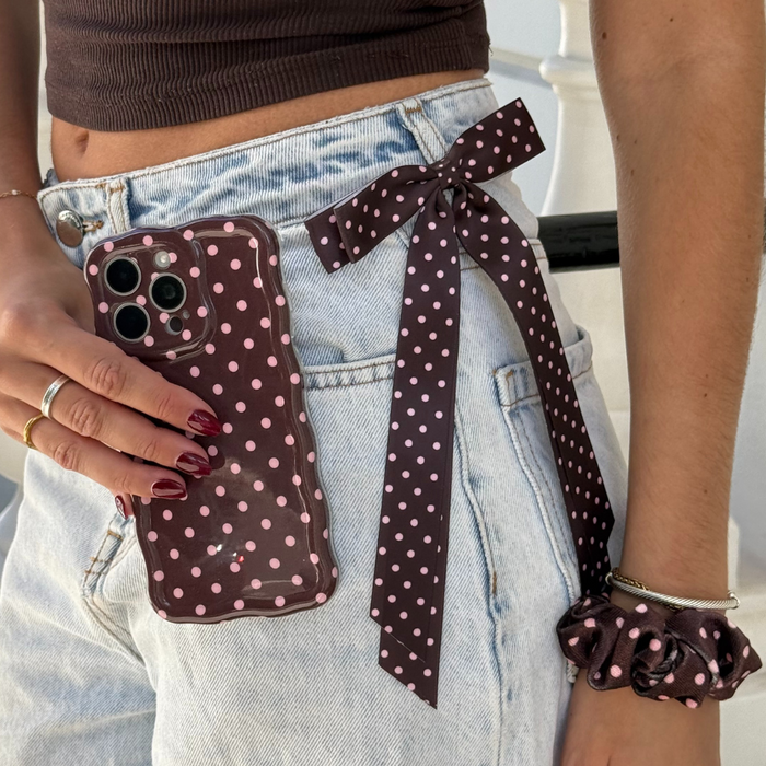Person in jeans holds a brown polka dot ribbon bow clip, polka dot phone, scrunchie, and has dark red nails.