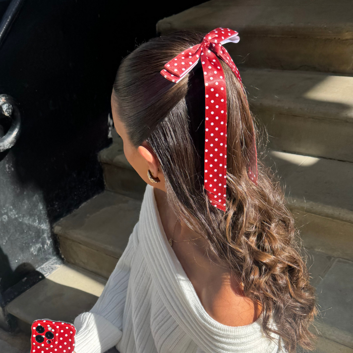 Woman with wavy hair wears a burgundy polka dot ribbon bow clip, seated on sunlit stairs.