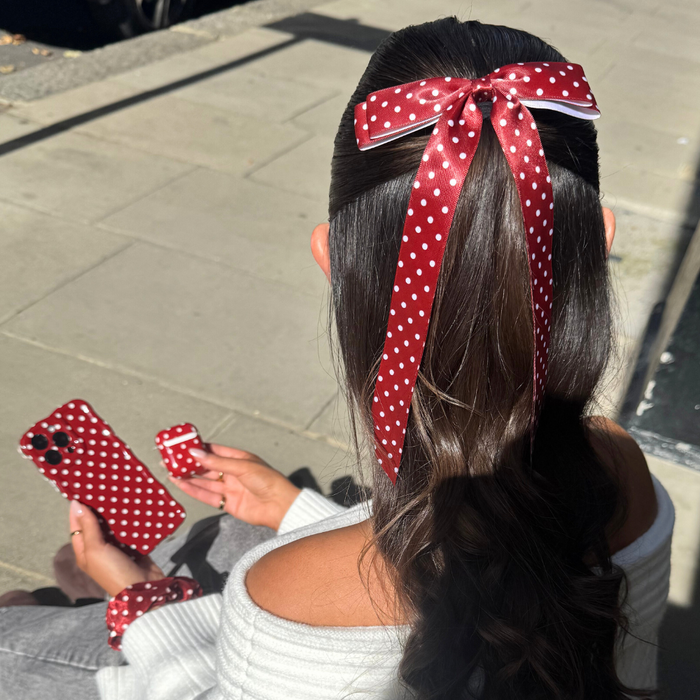 Woman with wavy hair wears a burgundy polka dot ribbon bow clip, holding matching burgundy phone and AirPods cases.