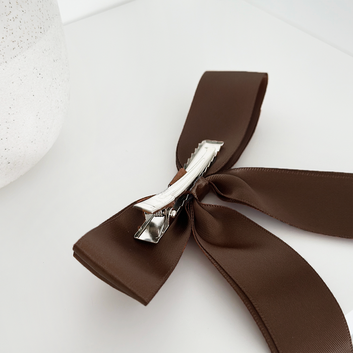 Chocolate brown ribbon bow hair clip with a silver clasp on a white surface next to a textured item.