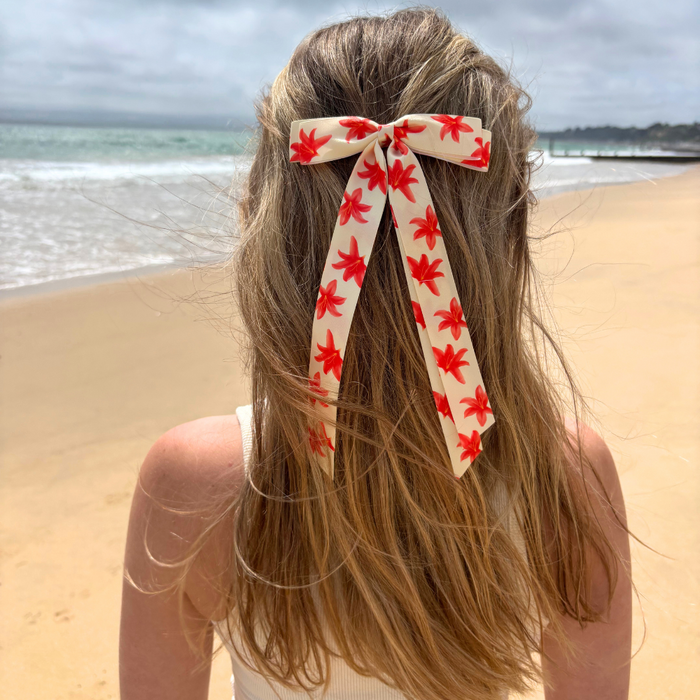 Woman on a sandy beach wears Coconut Lanes Ribbon Bow Clip featuring an Ibiza Lily print, facing the ocean.