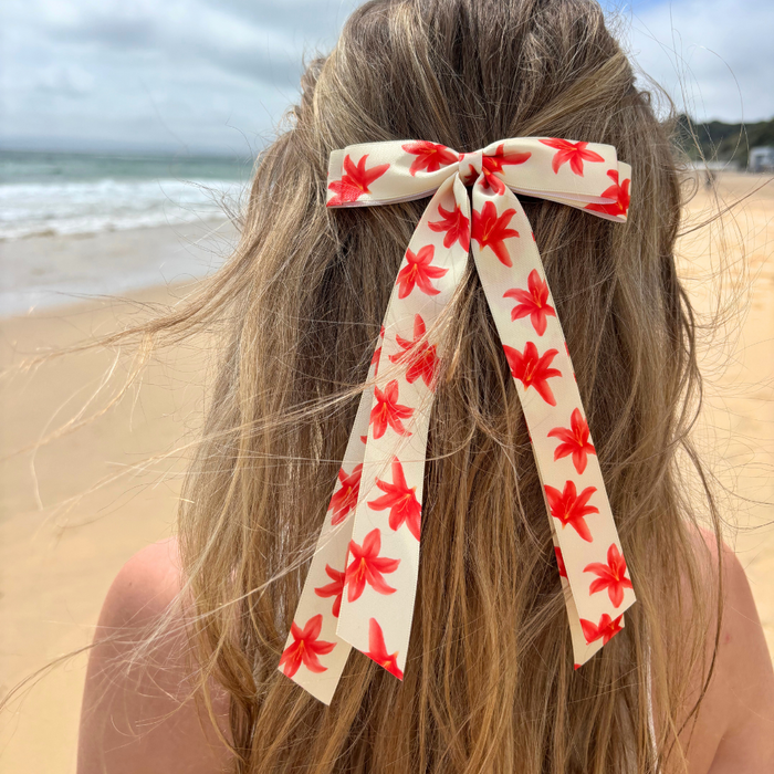 Long-haired person wearing a white ribbon bow hair clip on a sandy beach with the ocean in the background.