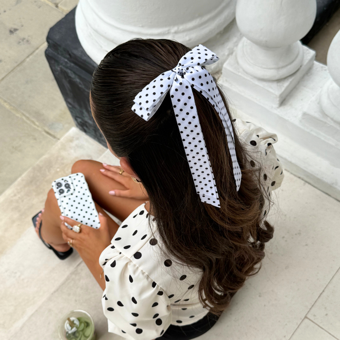 Woman with wavy hair wears a white polka dot ribbon bow clip, sitting on steps in a matching polka dot top.