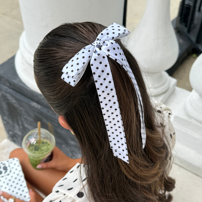 Woman with long brown hair wears a white polka dot ribbon bow clip, holding a green drink outdoors.