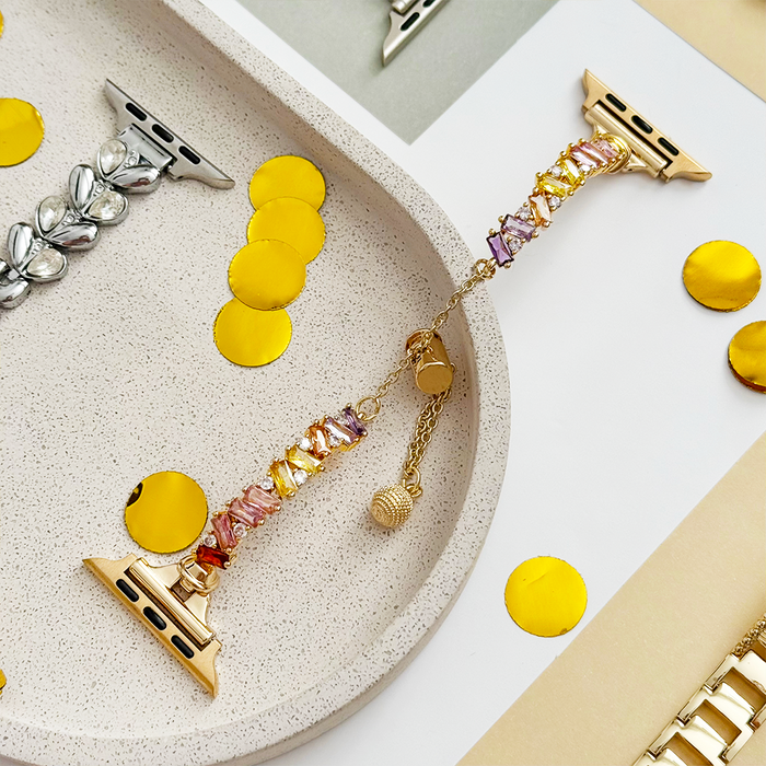 Gold crystal Apple Watch strap displayed on a tray with gold confetti and other watch bands nearby.