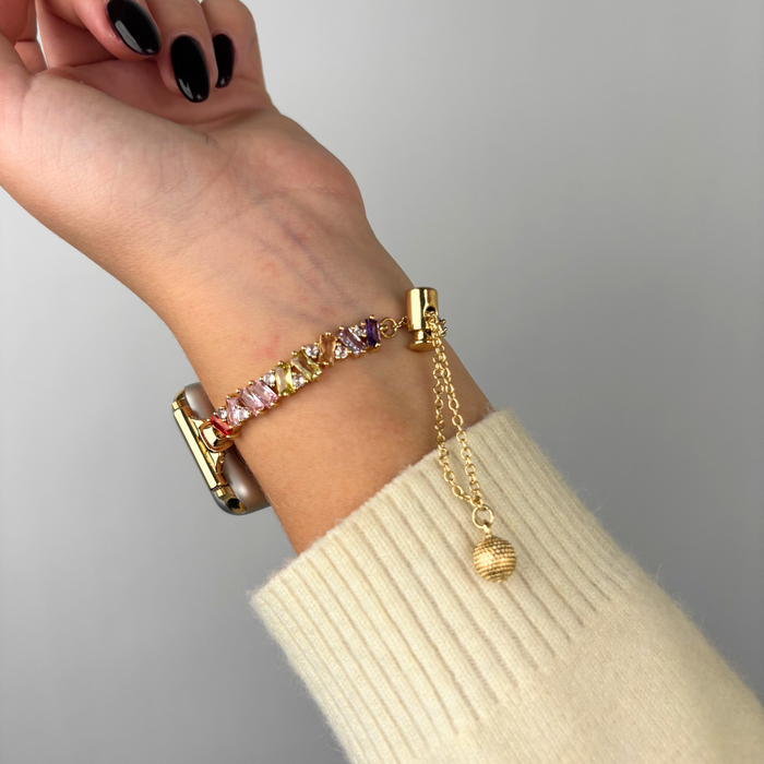 Gold Coconut Lane Apple Watch strap with colorful gems and a dangling charm on a wrist.