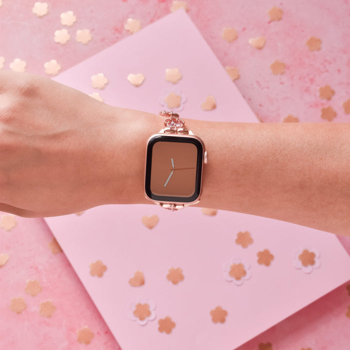 A person's wrist adorned with a stylish smartwatch, featuring the Rose Gold Flower Bracelet Apple Watch Strap from Coconut Lane. The background is a pink surface scattered with small heart-shaped and flower-shaped confetti, adding a festive touch.