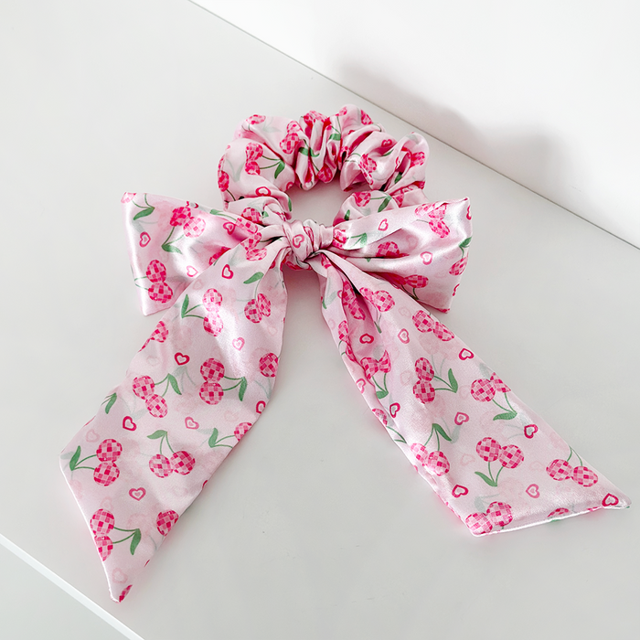 Pink satin bow scrunchie with heart pattern, displayed on a white surface.