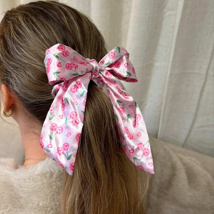 Woman tying her hair with a satin bow scrunchie featuring a cherry pattern, standing by beige curtains.