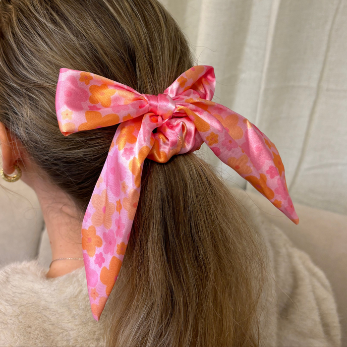 Satin bow scrunchie in pink and orange bloom pattern securing a low ponytail on light brown hair.