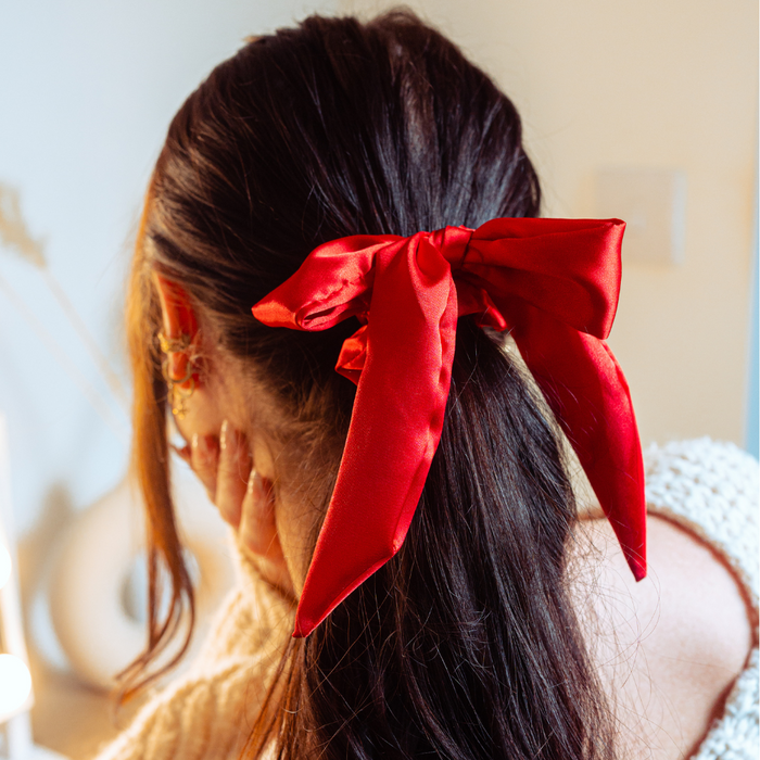 Woman with long brown hair wears a rose red satin bow scrunchie, shown from behind with one hand on her cheek.