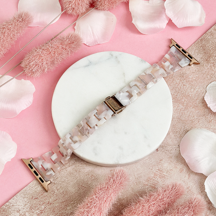 Slim Luxe Rose Apple Watch strap on a marble stand, surrounded by pink flowers and petals.