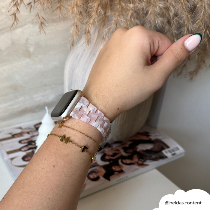 Wrist with a pink Slim Luxe Apple Watch strap and gold bracelets near a vase of dried grasses.