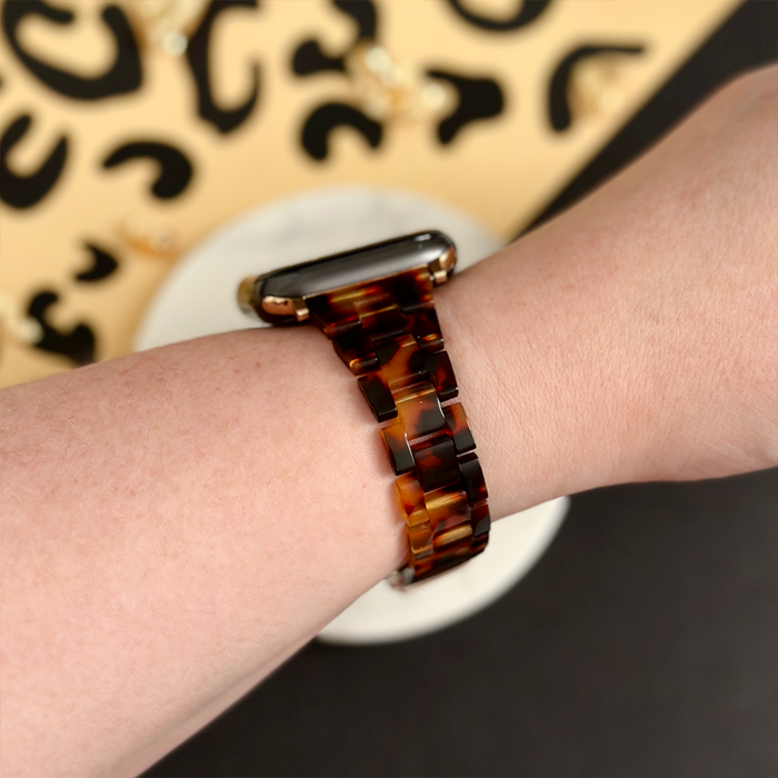 Wrist with Coconut Lane Slim Luxe tortoiseshell Apple Watch strap, thin and glossy, shown against a patterned background.
