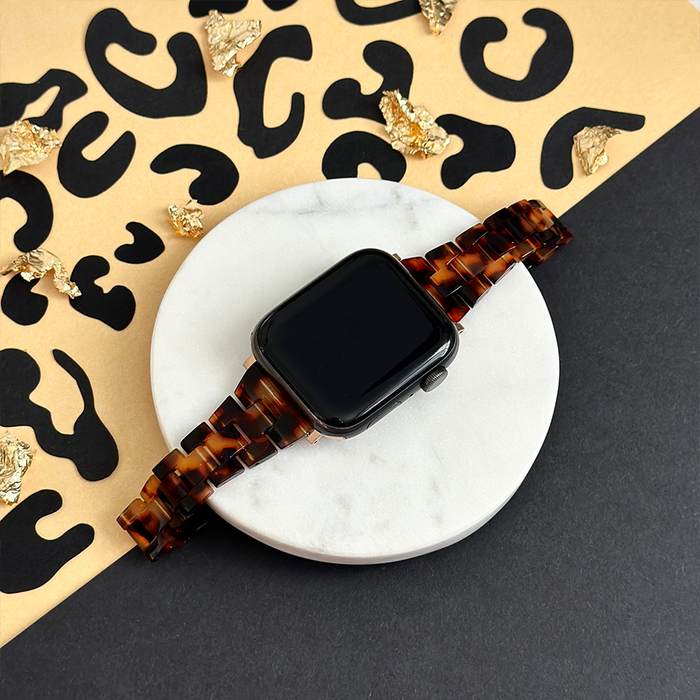 Slim Luxe tortoiseshell Apple Watch strap with gold hardware on white marble, leopard-print background visible.