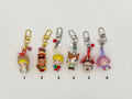 Six numbered character keychains on a white background. Features: (1) blonde girl in a bear hood with gold clasp, bell and star charms, (2) brown character in a red marching band uniform with gold clasp, (3) blonde boy holding a ball with silver clasp and bead charms, (4) freckled girl in brown outfit with silver clasp, (5) hedgehog in a red house hat with clock detail, (6) pink boy in lavender outfit with crystal charms.