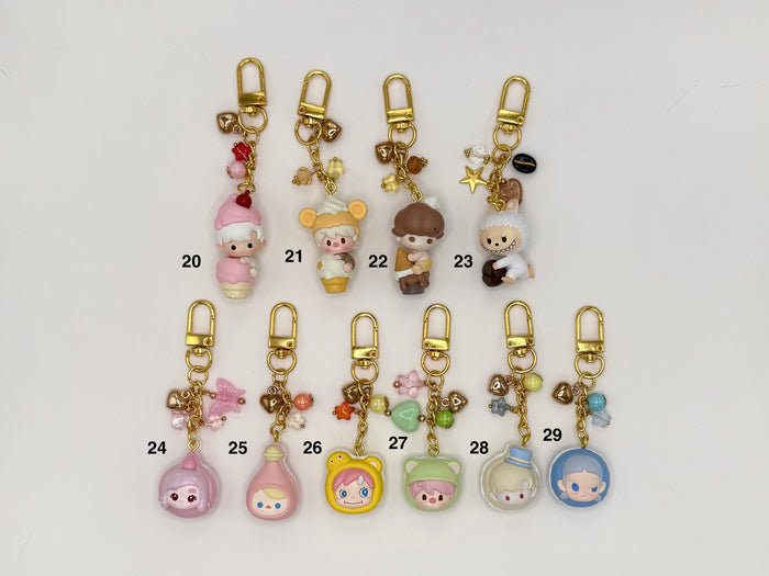 Ten numbered kawaii character keychains with gold clasps on a white background. Includes a pink elf girl (20), cream mouse in ice cream cone costume (21), mushroom-cap boy (22), white wolf with star charms (23), pink ghost figure (24), pink potion bottle (25), yellow bear face (26), green hooded character (27), and two clear dome figures with characters inside (28–29), all with decorative bead and chain accents.
