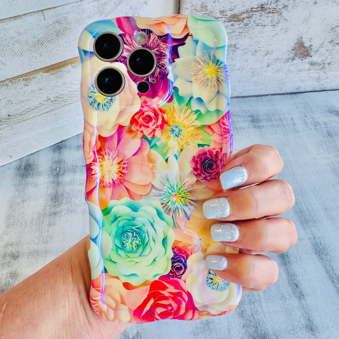 Hand holding a vibrant floral iPhone case featuring roses, daisies, and colorful blooms in pink, teal, and yellow with wavy edges.