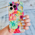 Hand holding a vibrant floral iPhone case featuring roses, daisies, and colorful blooms in pink, teal, and yellow with wavy edges.