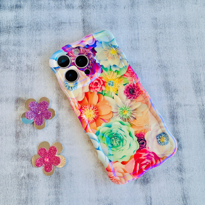Hand holding a vibrant floral iPhone case featuring roses, daisies, and colorful blooms in pink, teal, and yellow with wavy edges.