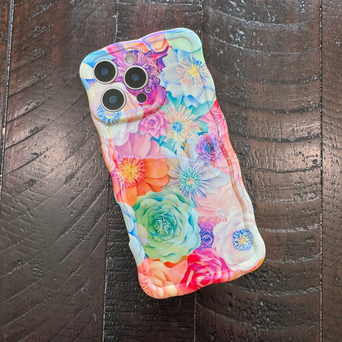 Hand holding a vibrant floral iPhone case featuring roses, daisies, and colorful blooms in pink, teal, and yellow with wavy edges.