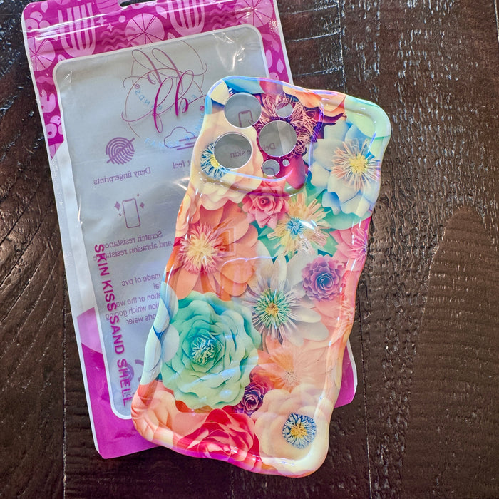 Hand holding a vibrant floral iPhone case featuring roses, daisies, and colorful blooms in pink, teal, and yellow with wavy edges.