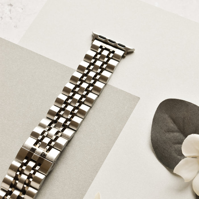 The Coconut Lane Stainless Steel Apple Watch Strap - Black & Silver, featuring a sleek, segmented design, is laid out on a light-colored surface. Nearby, a dark gray leaf and a small white flower add a touch of natural contrast to the modern accessory.