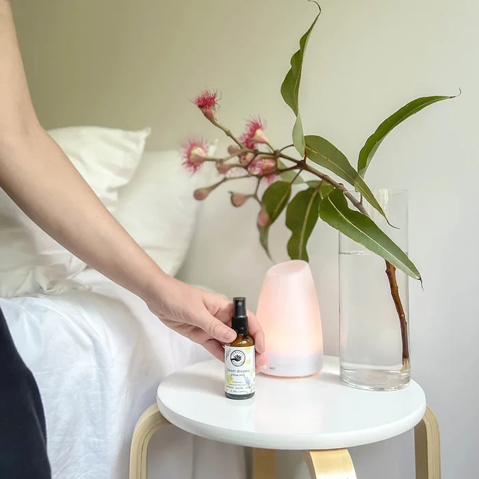 Hand holding a bottle of sweet dreams pillow mist next to a diffuser and a plant on a small table.