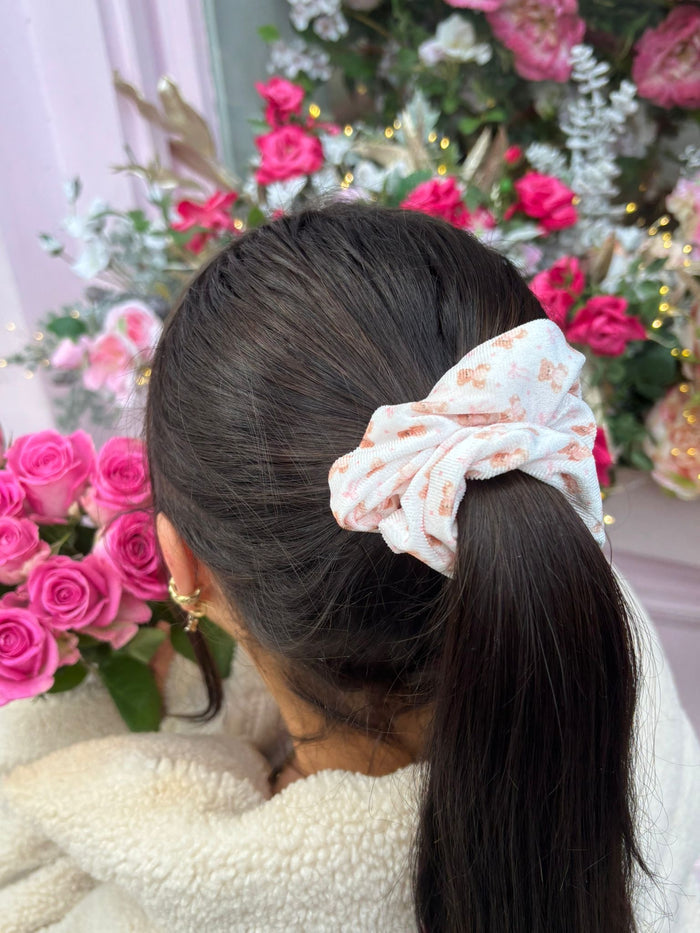 Woman with dark hair in a ponytail wears a fluffy beige scrunchie, holding pink roses with flowers in the background.