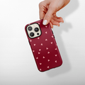 Hand holding a Coconut Lane MagSafe® phone case with pink hearts on a maroon background, white backdrop.