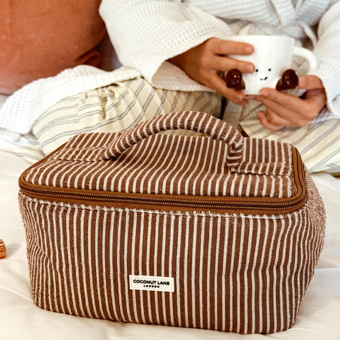 Chestnut striped vanity case by Coconut Lane on a bed, with pajama-clad person holding a mug in the background.