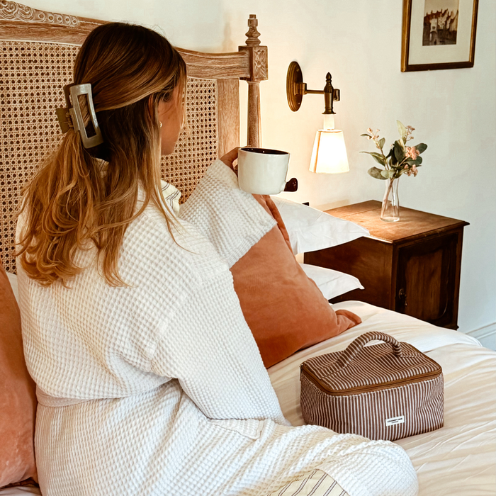 Woman in a robe on a bed with peach pillows, holding a mug beside a Coconut Lane chestnut-striped vanity case.