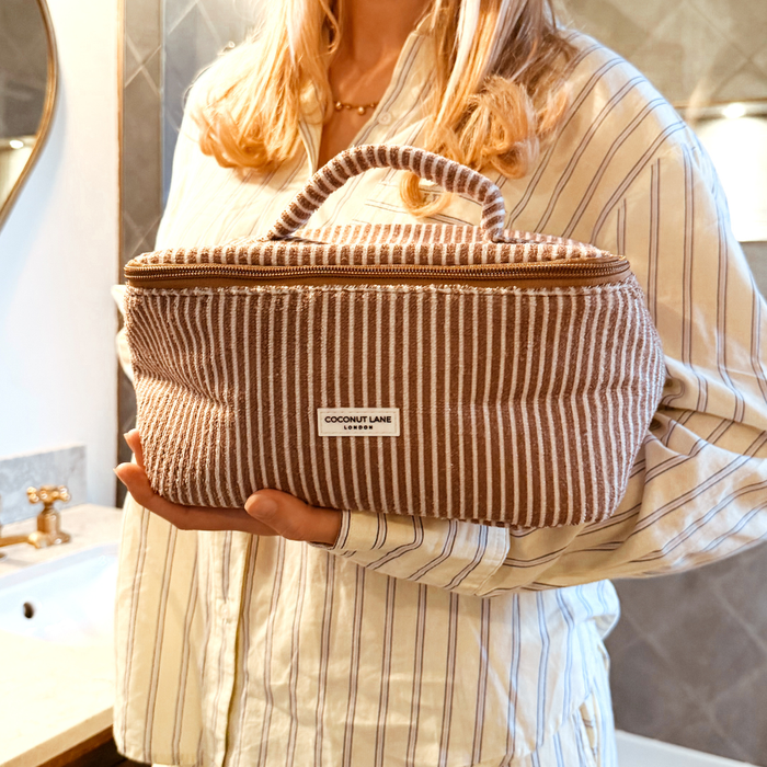 Woman in striped pajamas holds a chestnut striped Coconut Lane vanity case in a bathroom.