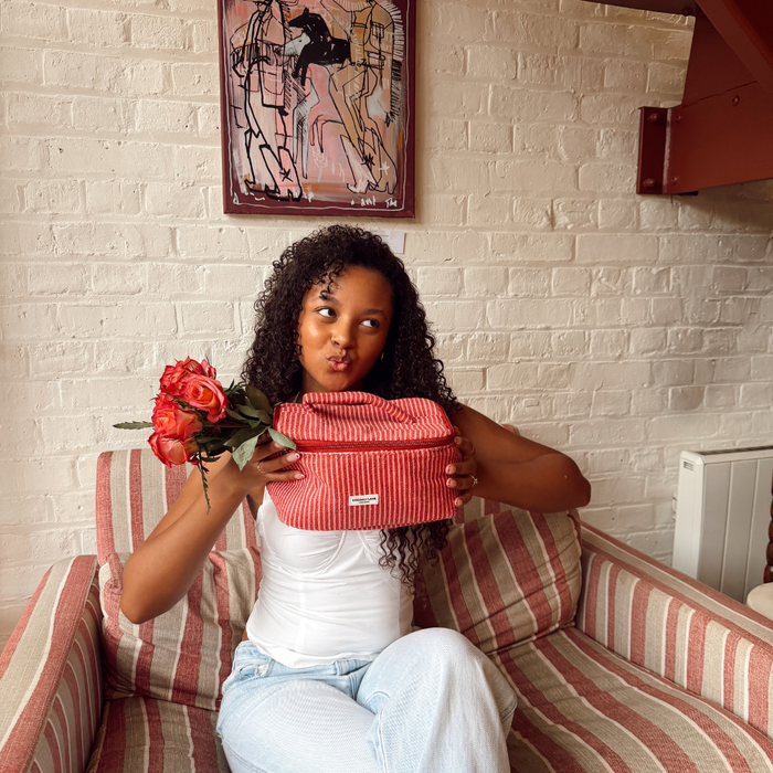 Woman seated on striped chair beside red flowers, red box, and striped Coconut Lane vanity case by an art-filled wall.
