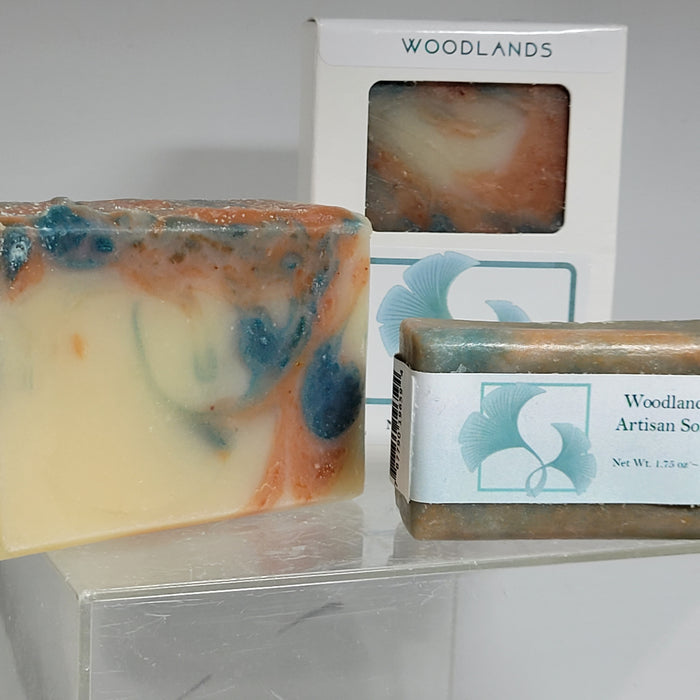 A bar of soap with swirls of blue, orange, and beige, alongside a packaged soap bar with a label reading 'Woodlands Artisan Soap'.